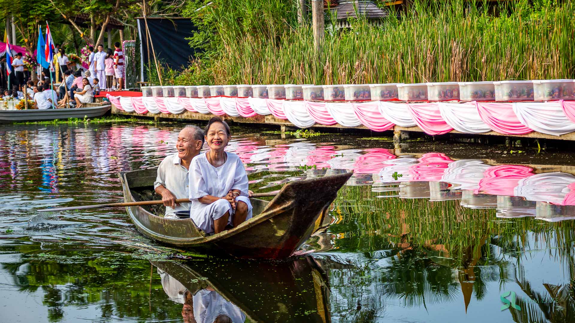 A Thai couple smile while paddling a wooden paddle boat to participate in the almsgiving ceremony on boat near the Ta Kha Floating Market